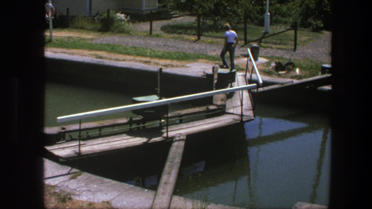 Man walks along the canal edge near a narrow bridge and railing.