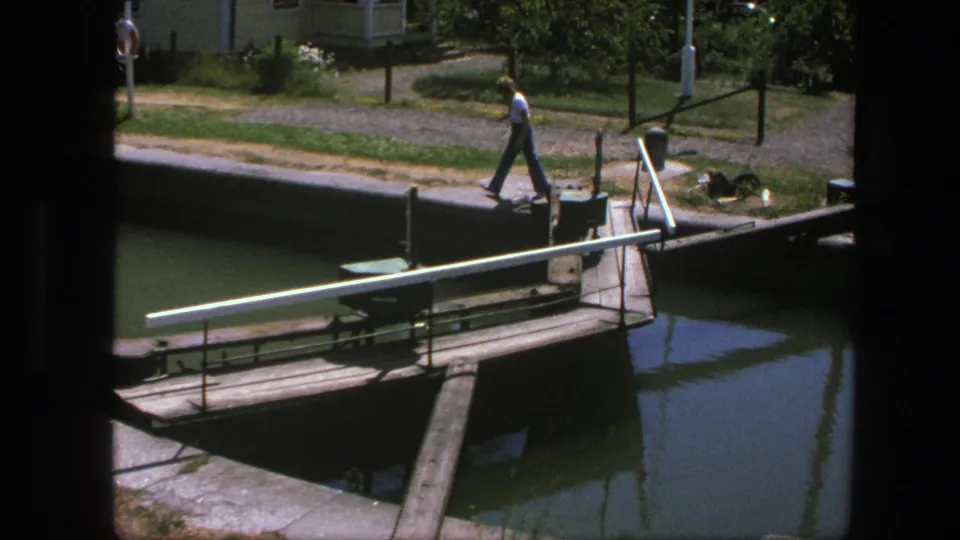 The man continues past the small bridge in Copenhagen, framed by canal water, railings, grass, and a dark foreground edge.
