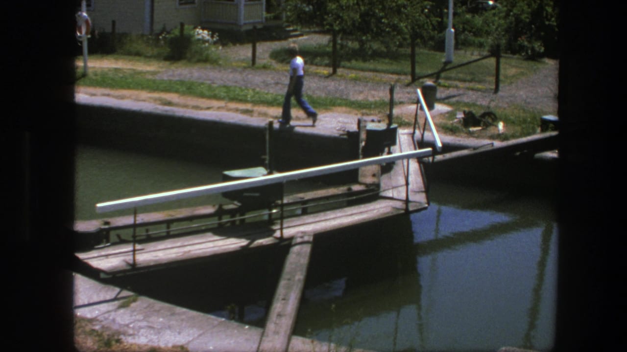 1975 Copenhagen home-movie frame showing a man on a footpath beside canal water and a wooden lock mechanism.