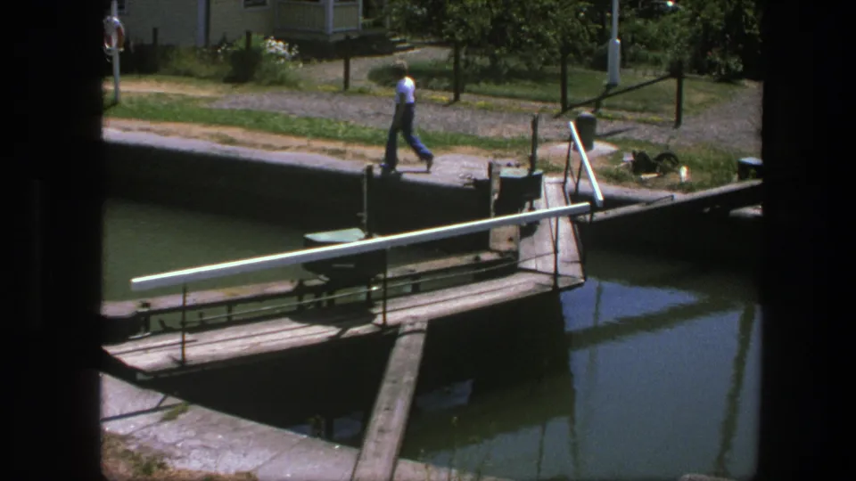 1975 Copenhagen home-movie frame showing a man on a footpath beside canal water and a wooden lock mechanism.
