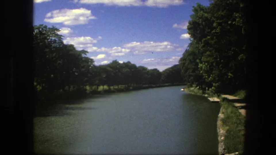 1975 Copenhagen canal view with calm water, dense green trees, stone bank, and scattered clouds above.