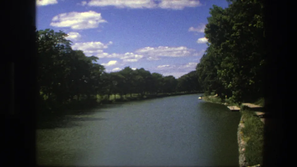 Copenhagen canal scene from 1975 with broad water, leafy banks, bright clouds, and a home-movie frame border.