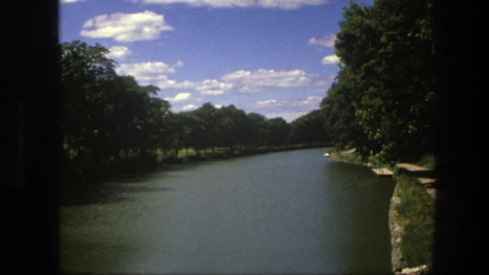 Late shot of the 1975 Copenhagen canal view, with green trees, calm water, stone edging, and bright summer clouds.
