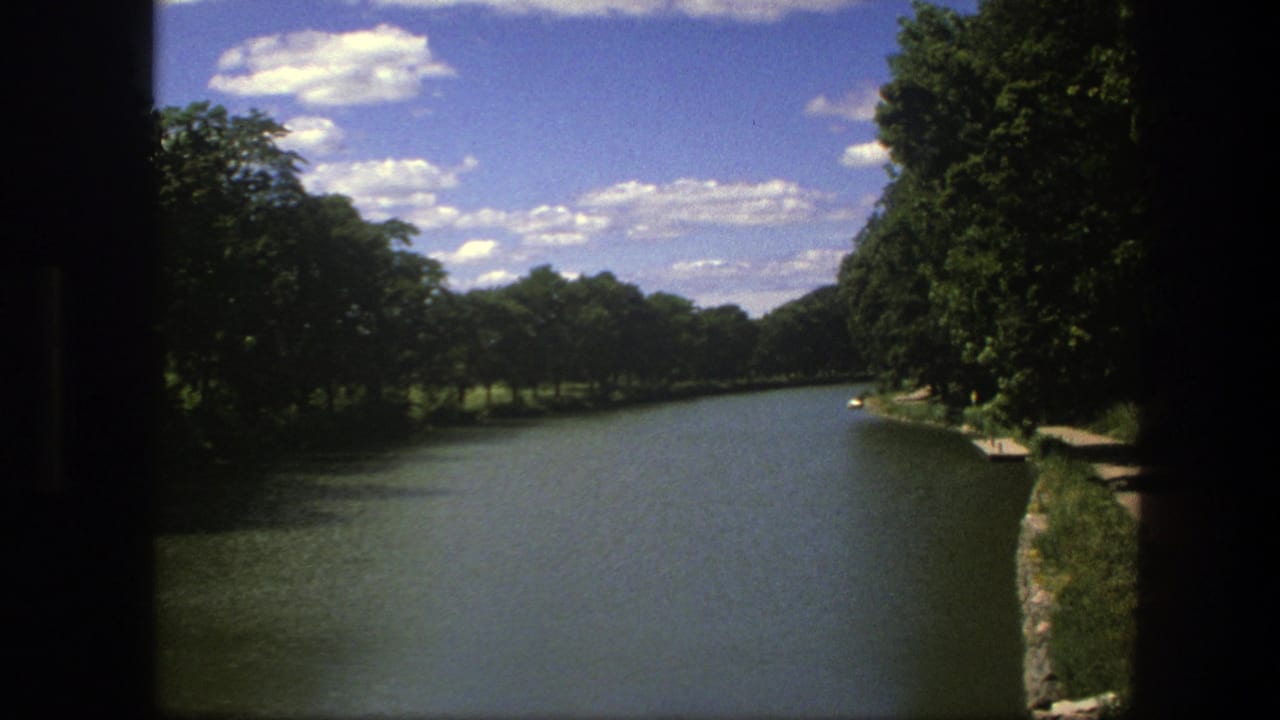 1975 Copenhagen 8mm frame showing a calm river between green tree banks beneath scattered white clouds.