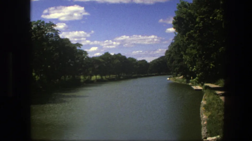1975 Copenhagen 8mm frame showing a calm river between green tree banks beneath scattered white clouds.