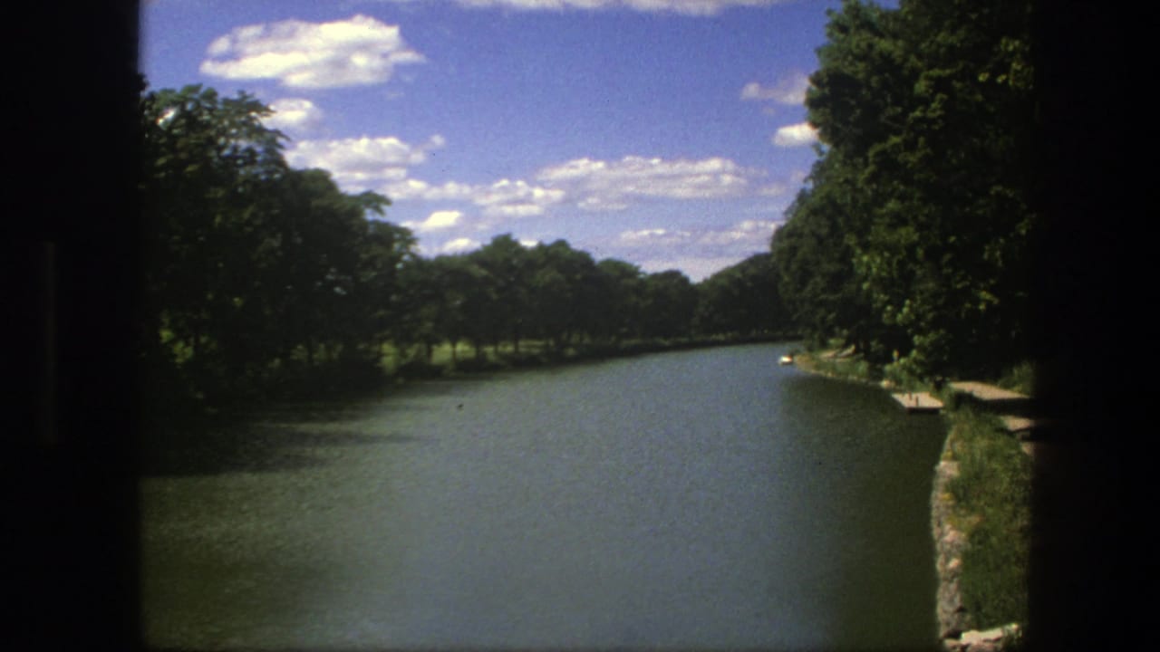 Calm waterway bordered by dense trees with a bright clouded sky above.