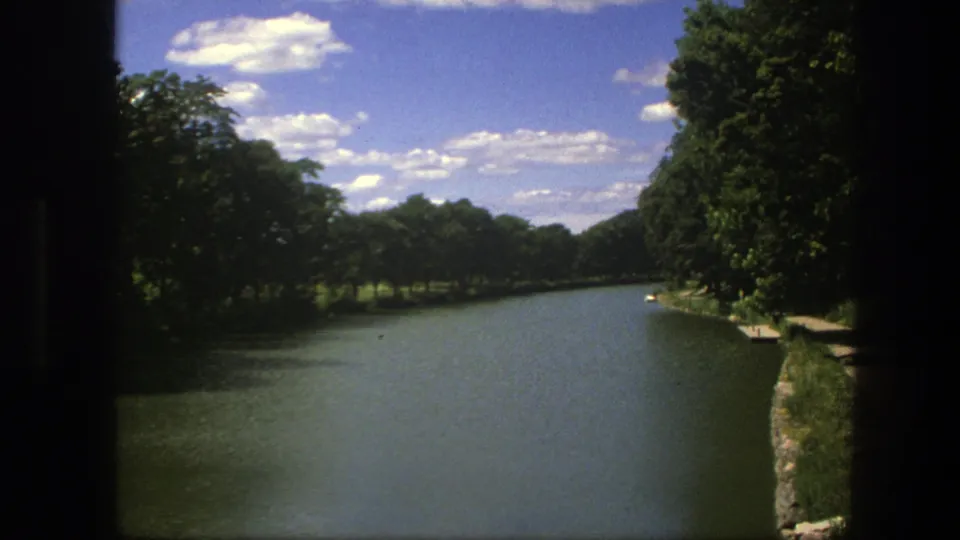 1975 Copenhagen home-movie frame of a quiet waterway with dense green trees and a small stone edge along the bank.