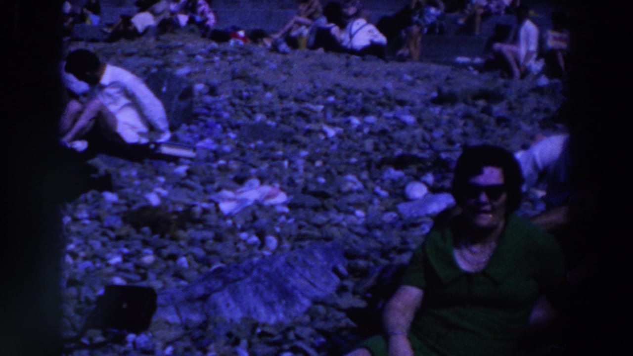 1961 Ireland home-movie frame showing a woman in a green dress seated on a pebbled beach, with scattered beachgoers in the background.