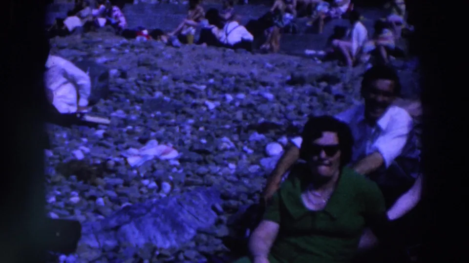 1961 Ireland beach footage frame with a seated woman in sunglasses in front and a man in a white shirt behind her on the rocky shore.