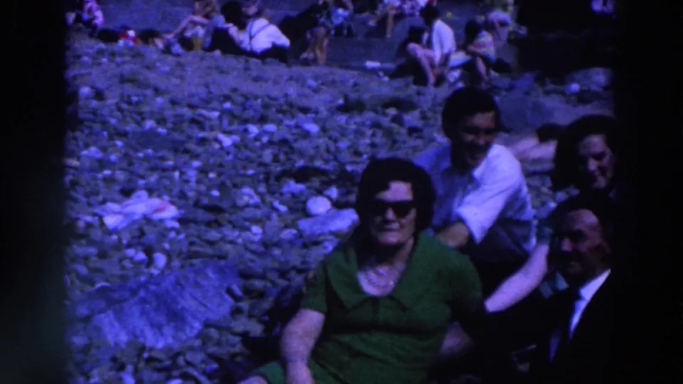 1961 Ireland home-movie frame showing several adults gathered on a pebbled beach, with the woman in green centered near the foreground.