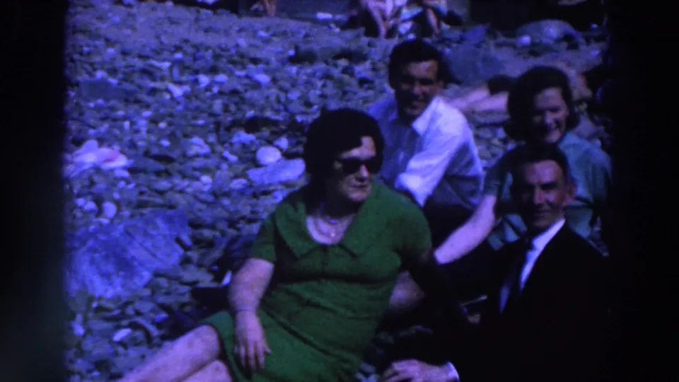 1961 Ireland frame showing four adults seated together on a pebbled beach, with visible faces and a blue-cast 8mm film look.