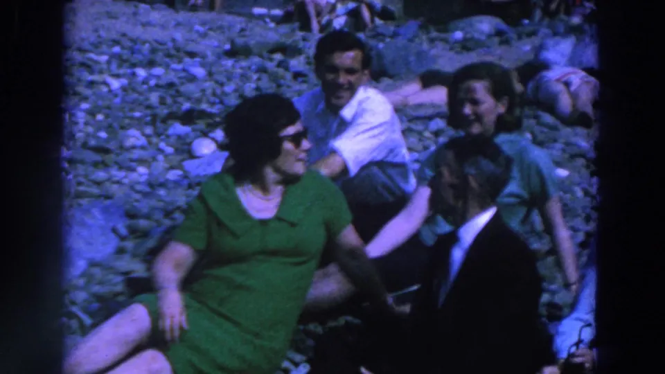 In 1961 Ireland, adults sit on rocky ground as the foreground woman in green turns toward a suited man beside her.