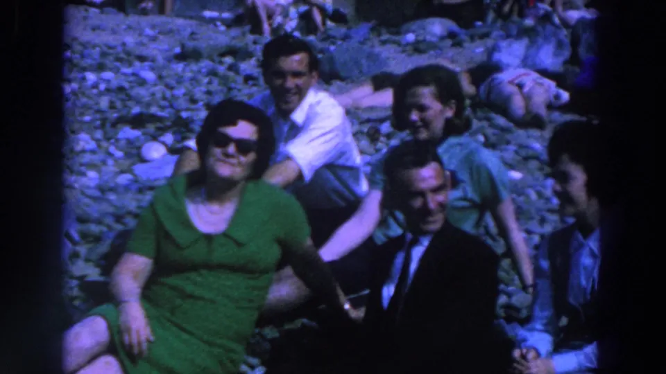 In 1961 Ireland home-movie footage, adults on gray rocks smile toward the camera as another figure enters at the edge.