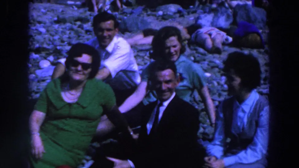 In 1961 Ireland, five adults sit closely on rocks in bright daylight, several smiling toward the home-movie camera.