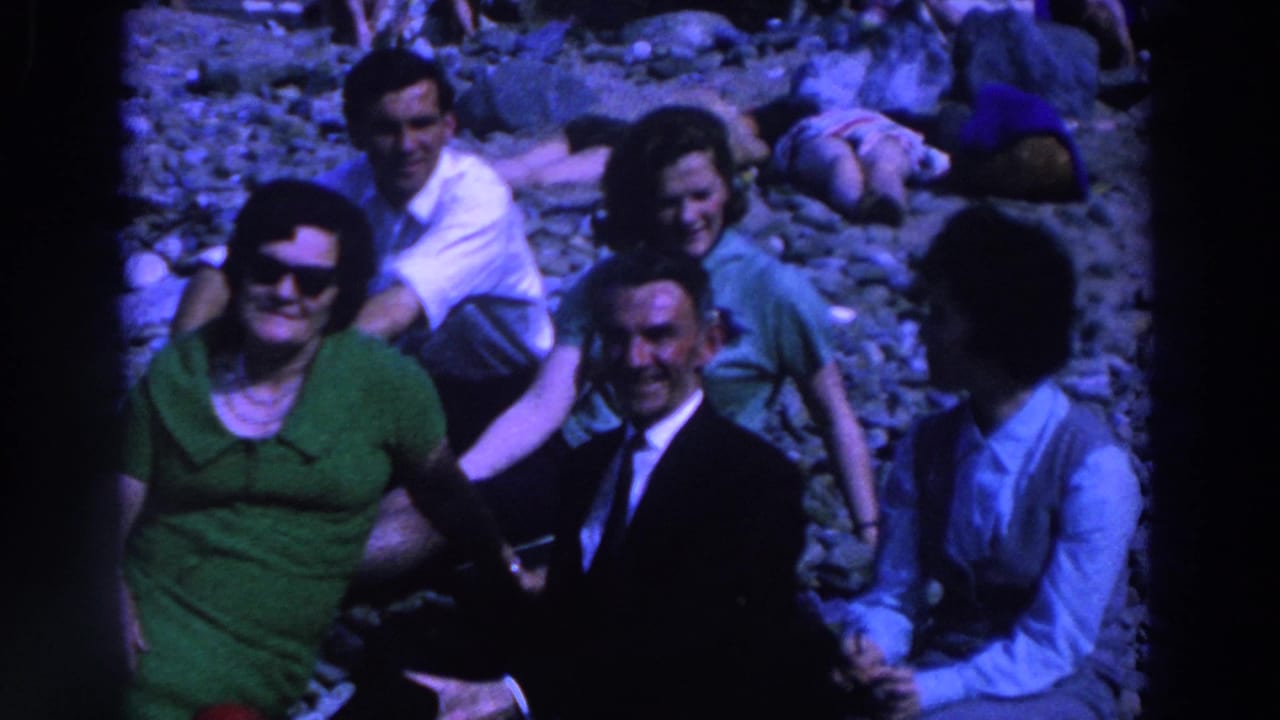 1961 Ireland home-movie frame showing adults relaxing on pebbles, including a woman in green sunglasses and a seated suited man.
