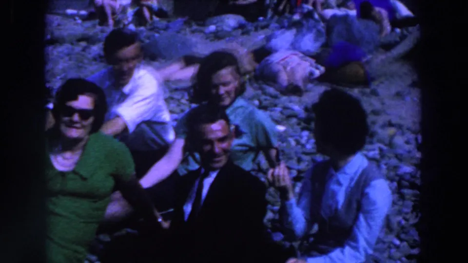 1961 Ireland 8mm frame of adults seated on a rocky shore, with a suited man smiling and others resting nearby.