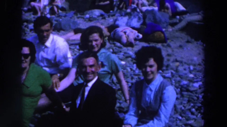 1961 Ireland home-movie view of seated adults on pebbles, with casual beachgoers and reclining figures behind them.