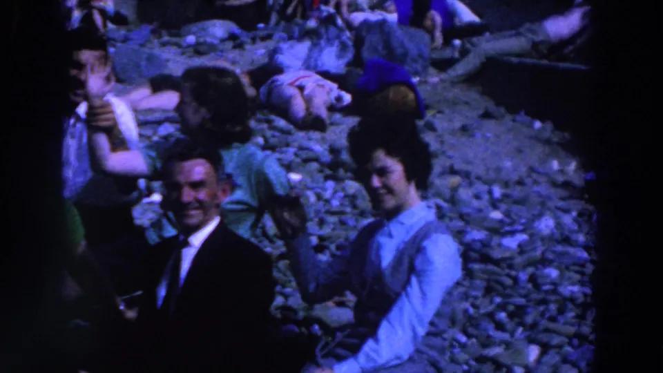 1961 Ireland 8mm frame showing adults seated together on a rocky beach, with background figures resting on the stones.