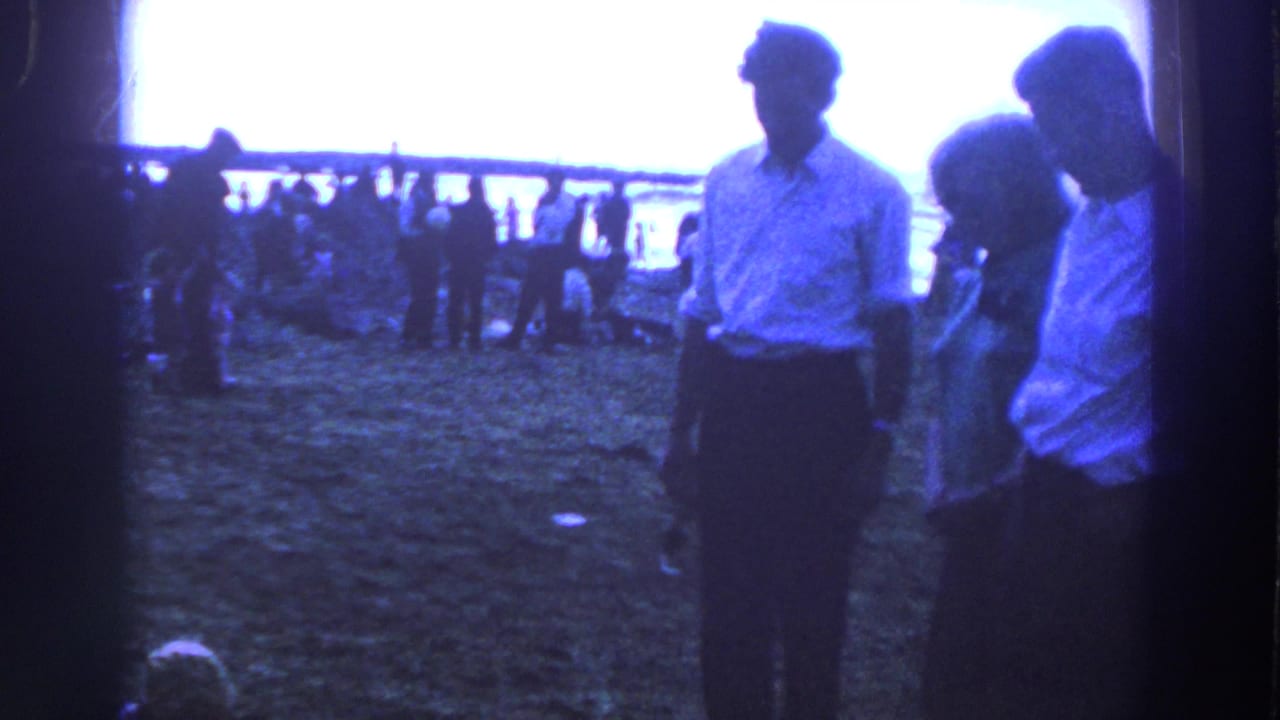 Three adults stand on grass near a railing with a crowd in the background.