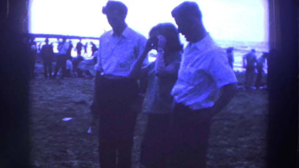 In the 1961 Ireland frame, the woman lifts a hand near her face while two men stand nearby on the grassy field.