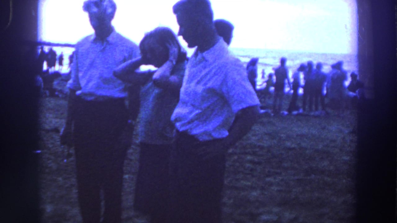 In 1961 Ireland, three adults stand on sandy beach ground while distant beachgoers gather near the shoreline.