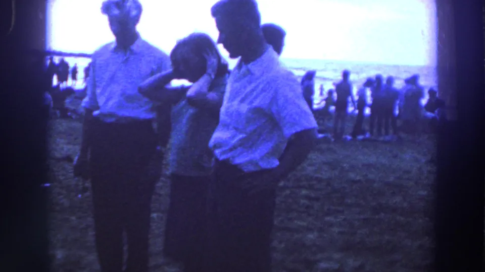In 1961 Ireland, three adults stand on sandy beach ground while distant beachgoers gather near the shoreline.