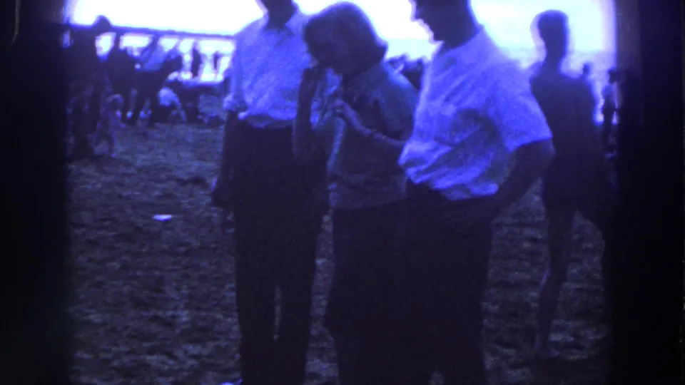 Adults in light shirts and a patterned dress stand on an Irish beach, with other visitors and a pier-like structure behind them.