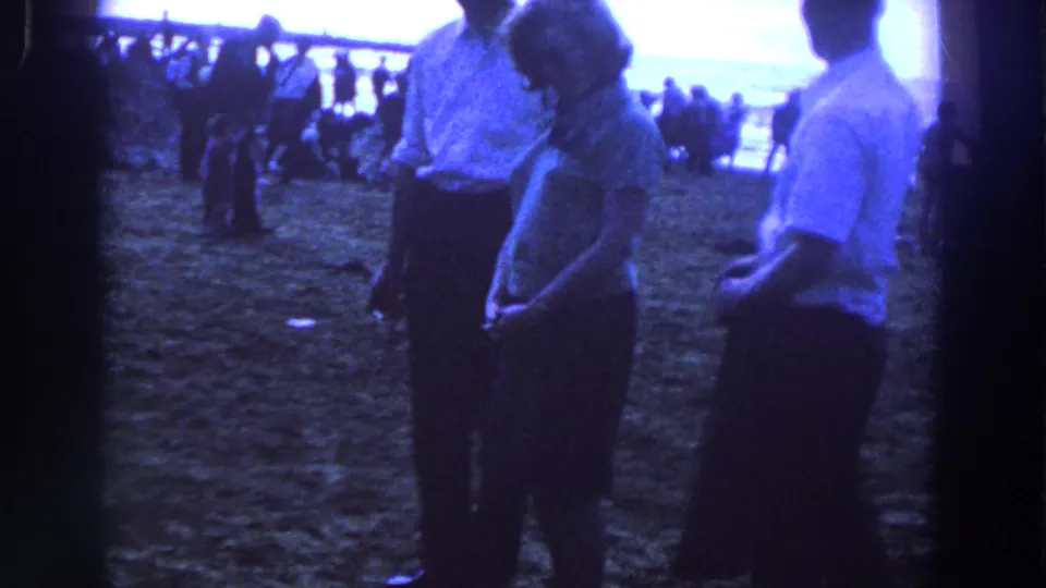 A woman in a patterned dress stands between two men on a 1961 Irish beach, with distant visitors near the water.