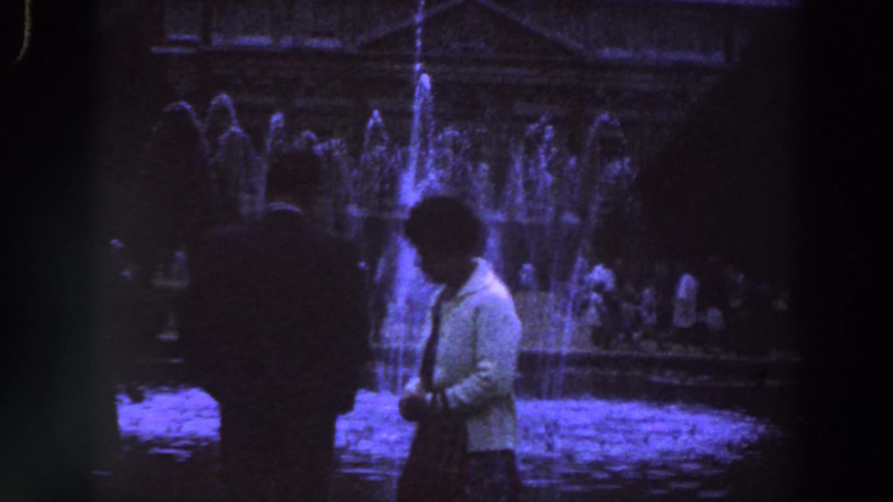 1961 Ireland home-movie frame with a man and woman walking before an illuminated public fountain at night.