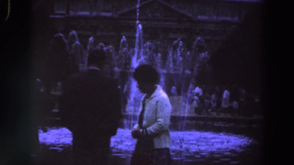1961 Ireland home-movie frame with a man and woman walking before an illuminated public fountain at night.