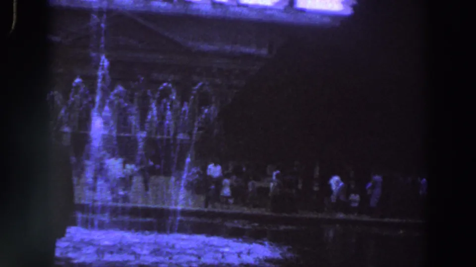 1961 Ireland frame of blue-lit fountain spray and distant visitors in a nighttime public setting.