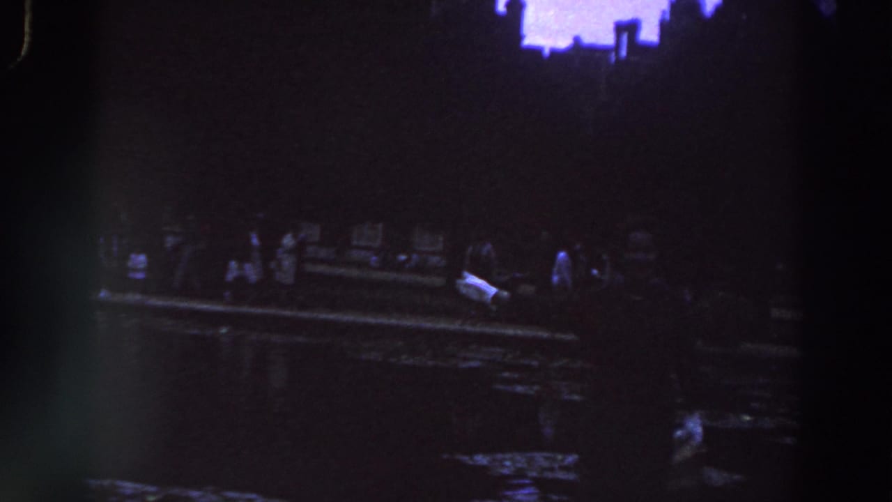 1961 Ireland home-movie frame showing dimly lit adults near a waterside edge, with dark reflections and a purple-blue cast.