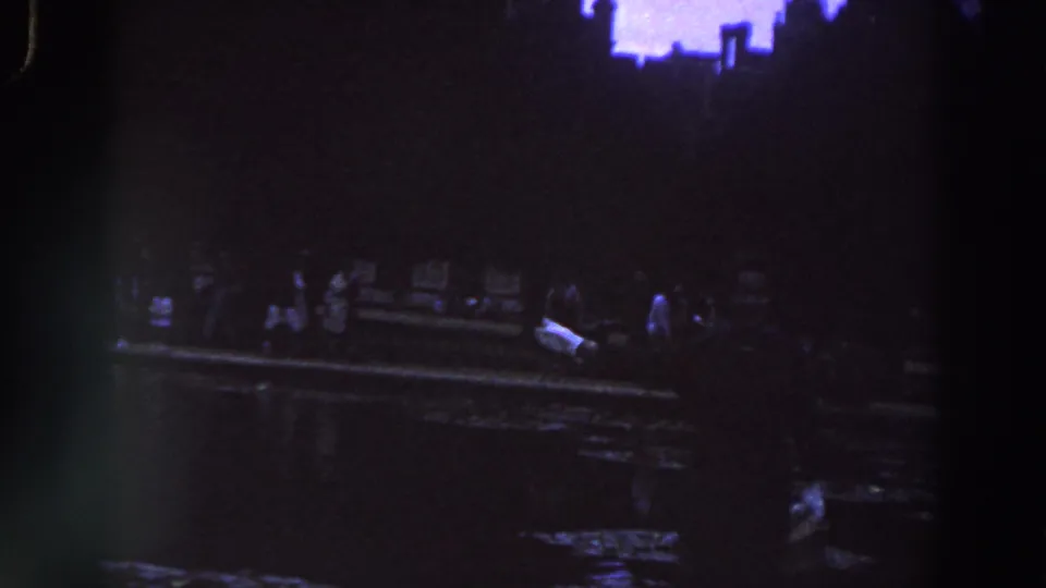 1961 Ireland home-movie frame showing dimly lit adults near a waterside edge, with dark reflections and a purple-blue cast.
