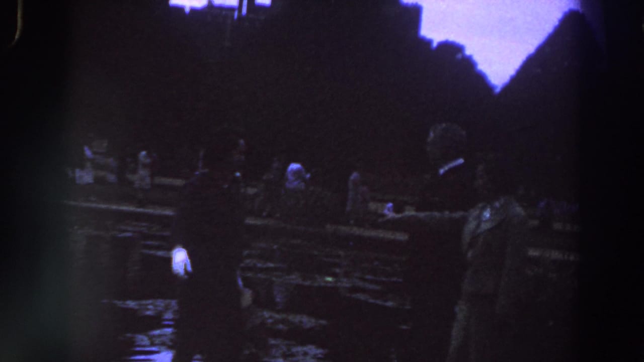 1961 Ireland home-movie frame showing adults in formal clothing near a wet park or garden area with dark silhouettes behind them.