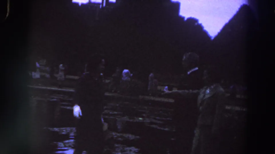 1961 Ireland home-movie frame showing adults in formal clothing near a wet park or garden area with dark silhouettes behind them.