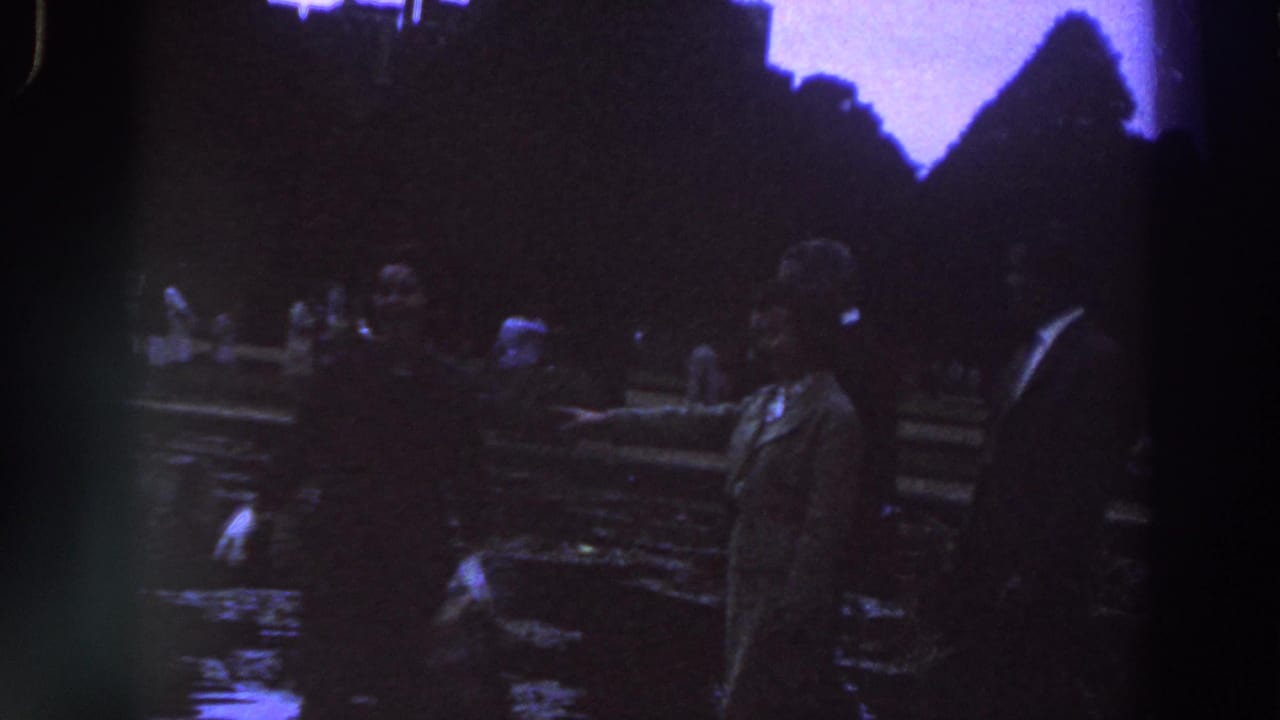 A woman in a coat and a suited man stand beside a wet garden walkway or pond edge.