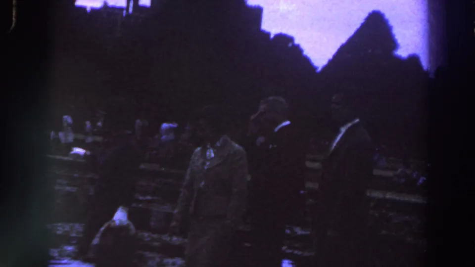 1961 Ireland home-movie frame of adults moving through a dim public garden or park, with wet ground and background visitors.