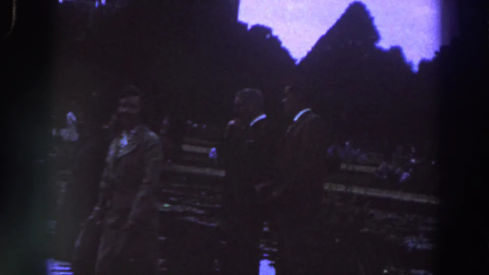 1961 Ireland frame showing a small group of adults in formal clothing moving through a public garden or park at low exposure.