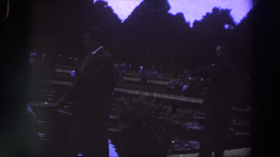 1961 Ireland 8mm frame with adults moving around a stone monument or statue base at dusk.