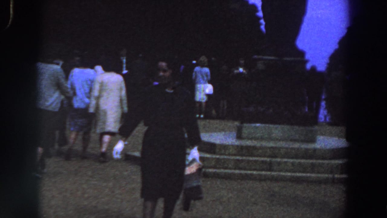 1961 Ireland home-movie frame showing a woman in a dark dress near stone steps as people in coats gather around a monument-like structure.