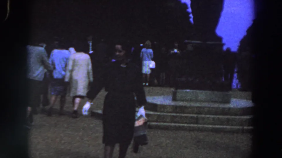 1961 Ireland home-movie frame showing a woman in a dark dress near stone steps as people in coats gather around a monument-like structure.
