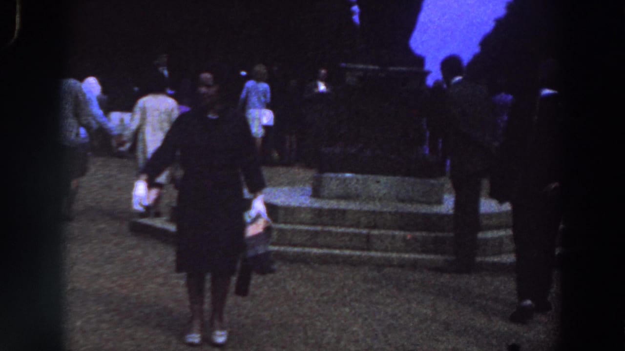 Woman in dark clothing stands near a crowd and stone monument base at night.