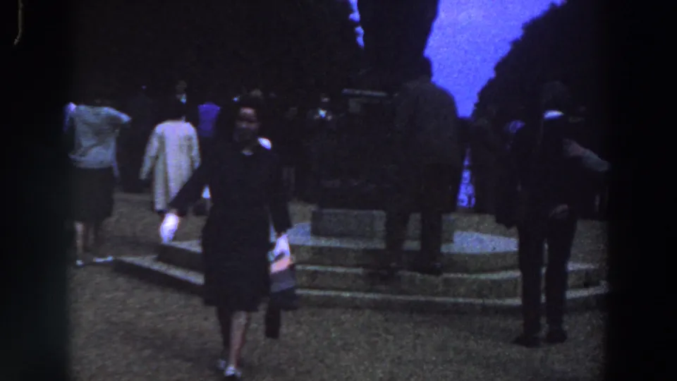 1961 Ireland home-movie frame with the woman now seen from behind as she moves among visitors near stone steps and a dark monument area.
