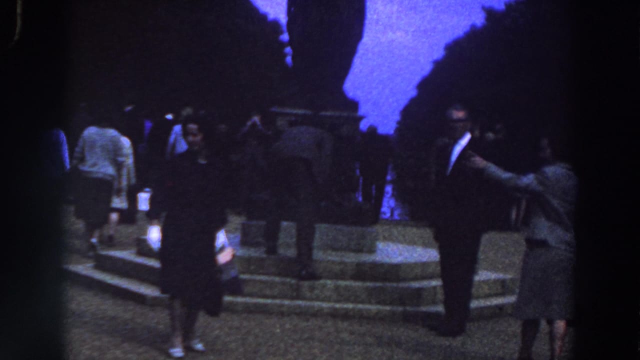 In Ireland home-movie footage from 1961, adults in suits and skirts stand near stone steps and a large dark monument under open sky.