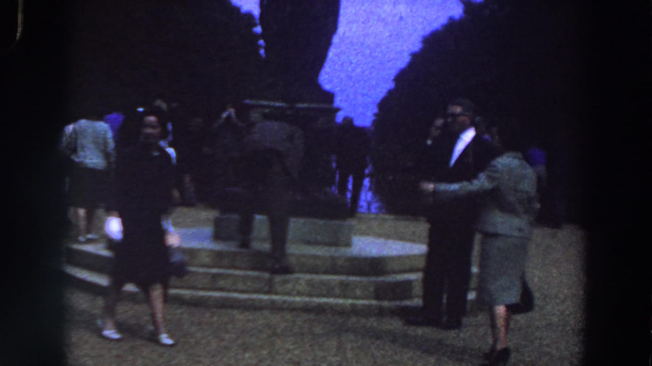 A woman walks across the foreground while adults stand beside a dark monument and stone platform.