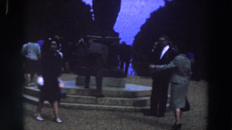 A 1961 Ireland home-movie frame shows a woman in a dark dress crossing near stone steps as other adults face a dark monument.