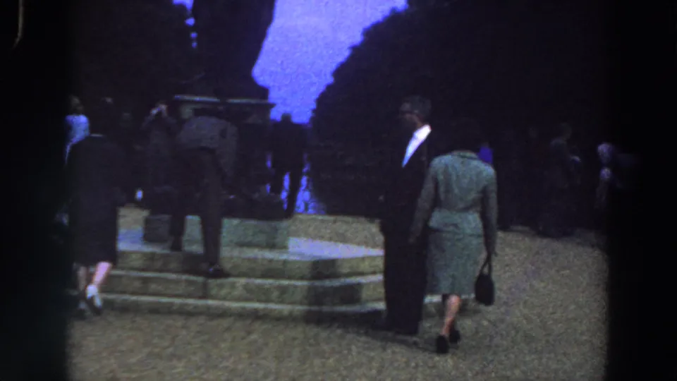 A woman in a grey skirt suit walks toward the dark monument in 1961 Ireland footage, with other adults near the stone steps.