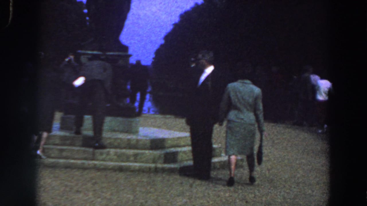 1961 Ireland 8mm frame showing a suited man and a woman in a green dress walking near stone steps in a park at dusk.