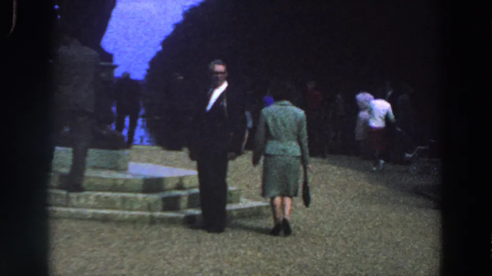 1961 Ireland park footage with a woman in a green dress walking away from camera near a suited man, stone steps, and distant visitors.
