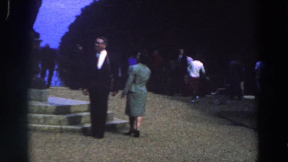 1961 Ireland 8mm still showing the couple beside stone steps and a dark monument, with park visitors and a stroller behind them.